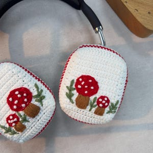 May include: White crocheted headphone covers with red and white mushroom embroidery. The mushrooms have green leaves and brown stems.