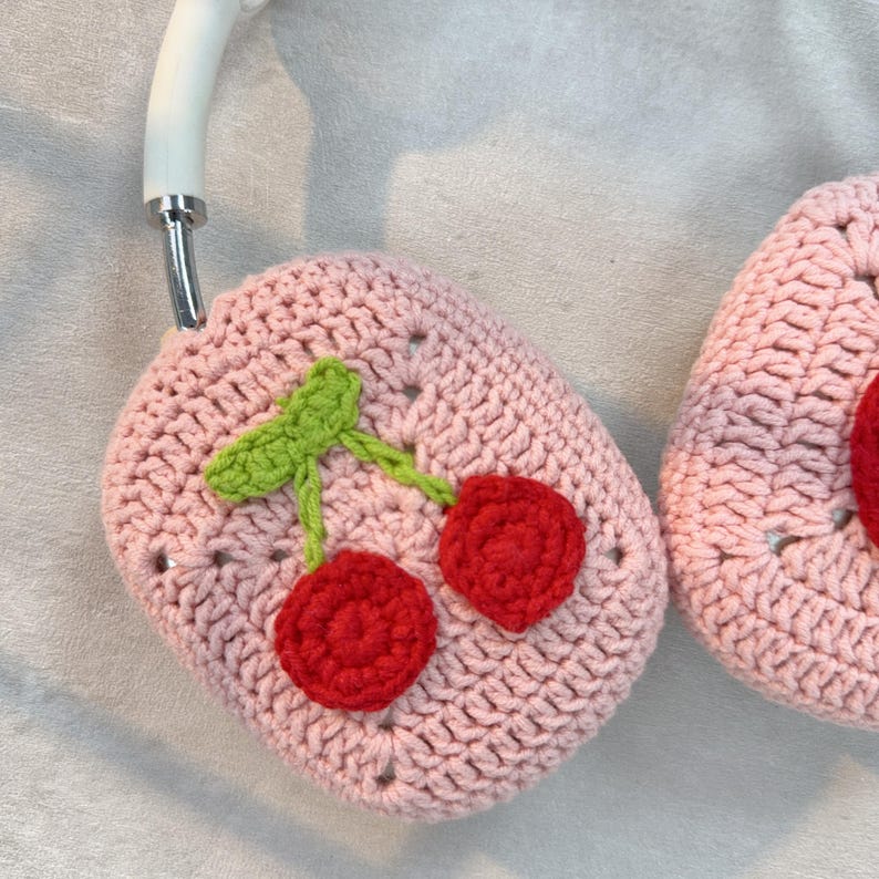May include: A pink crocheted case with a cherry design. The case has two cherries with green stems and leaves. The cherries are red and white.