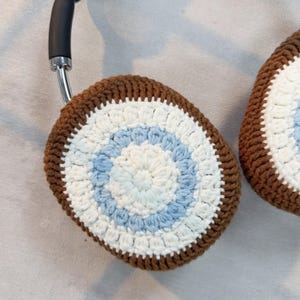 May include: A pair of brown, white, and blue crocheted earmuffs for headphones. The earmuffs are round and have a concentric circle pattern.