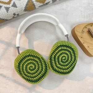 Puede incluir: Auriculares blancos con orejeras de ganchillo verde y verde oscuro. Las orejeras tienen un patr&oacute;n circular.