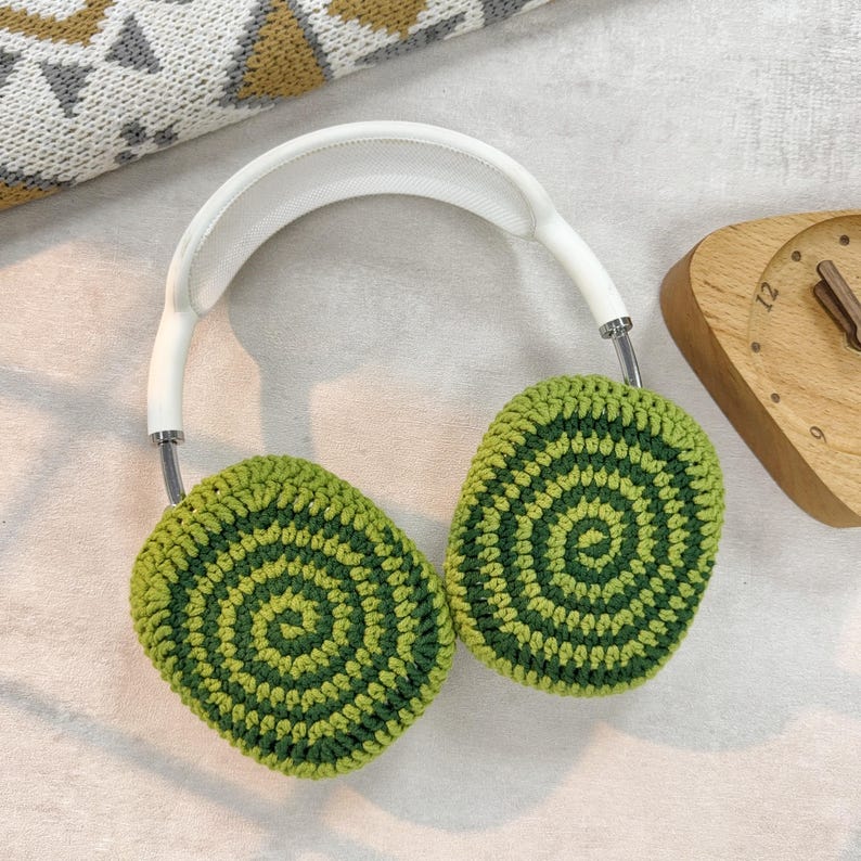 Puede incluir: Auriculares blancos con orejeras de ganchillo verde y verde oscuro. Las orejeras tienen un patr&oacute;n circular.