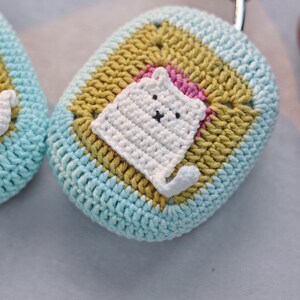 May include: A crocheted light blue and yellow case with a white crocheted cat design. The cat has a black nose and eyes.