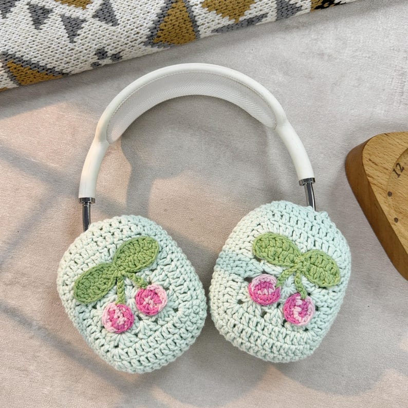 Pu&ograve; includere: Cuffie bianche con paraorecchie in crochet verde chiaro con un motivo a ciliegie. Le ciliegie sono rosa e rosse con foglie verdi.