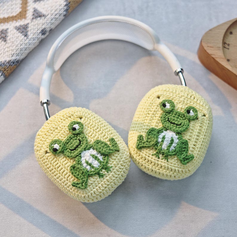 May include: White headphones with crocheted earmuffs featuring green frogs. The earmuffs are yellow with white accents.