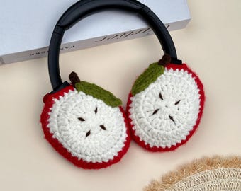 May include: Headphones with black headband and crochet ear covers. The ear covers are designed to look like apple slices, with red edges, white centers, green leaves, and brown stems. The headphones are resting on a light-colored surface.