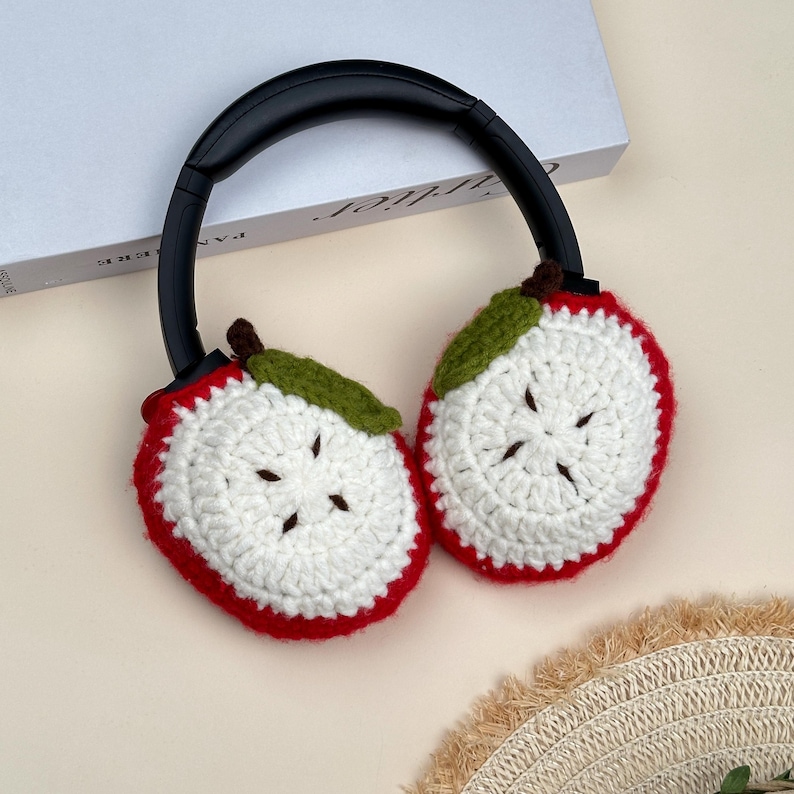 Puede incluir: Auriculares con diadema negra y orejeras de ganchillo. Las orejeras est&aacute;n dise&ntilde;adas como rodajas de manzana, con bordes rojos, centros blancos, hojas verdes y tallos marrones. Los auriculares est&aacute;n sobre una superficie clara.