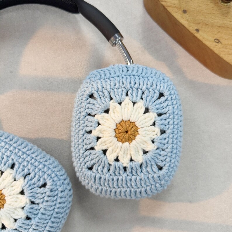 Puede incluir: Fundas para auriculares de crochet azul claro con un dise&ntilde;o de margarita blanca. La margarita tiene un centro marr&oacute;n.