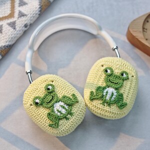 May include: A pair of crocheted earmuffs featuring a frog design. The earmuffs are yellow with green frogs and white accents. The earmuffs are attached to a white headband.