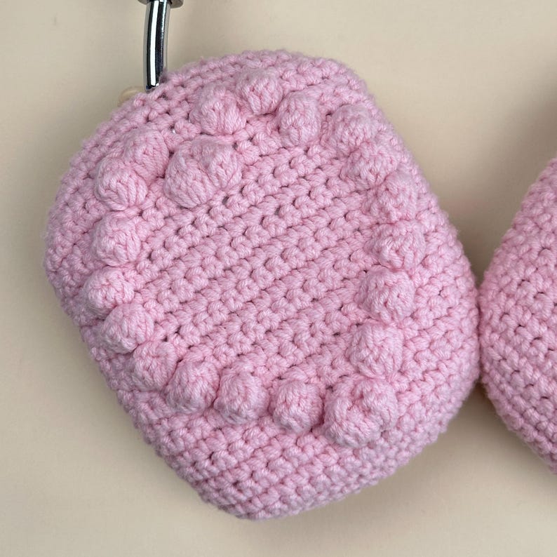 Op de afbeelding: Een paar lichtroze, handgemaakte gehaakte pannenlappen. De pannenlappen hebben een textuurontwerp met verhoogde, afgeronde bobbles. Het haakwerk is zichtbaar en de pannenlappen hangen aan een metalen haak.