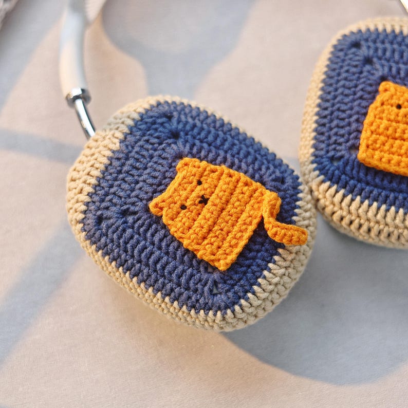May include: A pair of crocheted headphone covers with a cat design. The covers are blue and white with a yellow cat in the center.