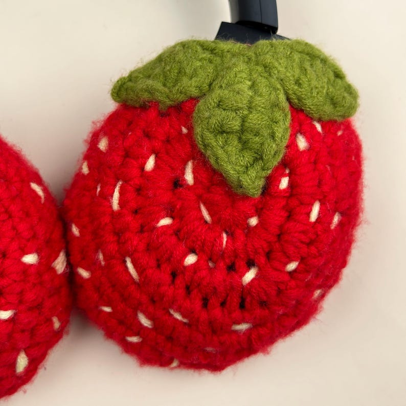 May include: Crocheted strawberry headphone covers. The red covers have white seed details and green leaf accents. The handmade covers are designed to fit over the ear cups of headphones. The strawberry design adds a playful touch to the headphones.