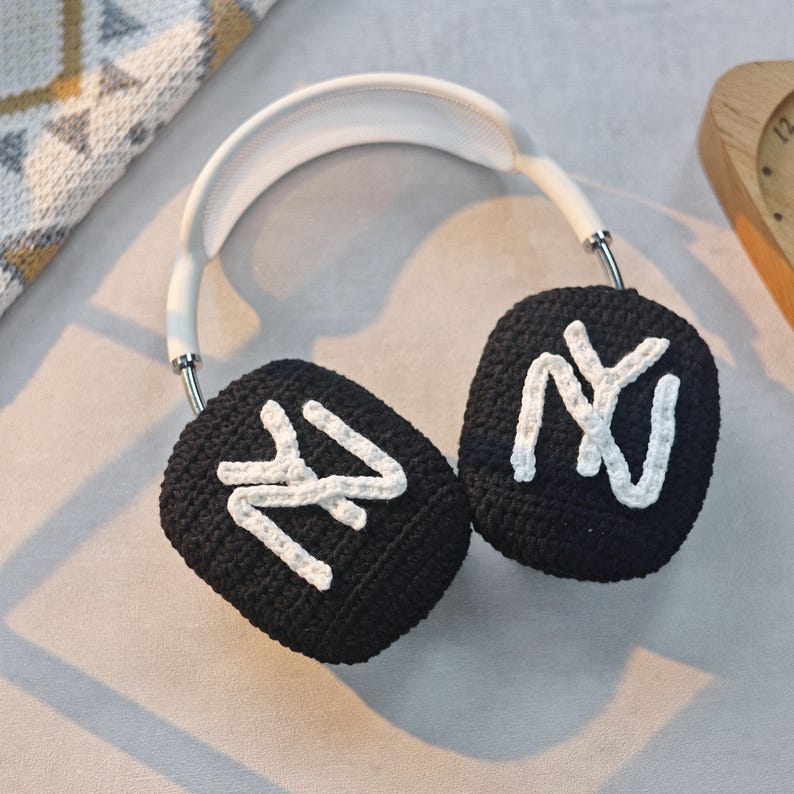 May include: White headphones with black crocheted earmuffs. The earmuffs have a white "NY" logo on each side.