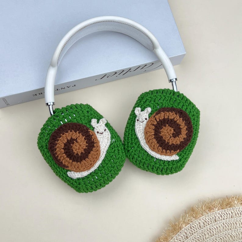 Pu&ograve; includere: Cuffie bianche con copriauricolari a uncinetto verdi, marroni e bianchi a forma di lumaca. I copriauricolari sono di un verde brillante con un disegno di lumaca marrone e bianco. L'archetto &egrave; bianco e gli accenti metallici sono argentati.