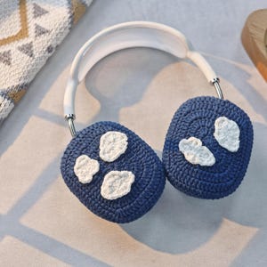 May include: Blue crocheted earmuffs with white crocheted cloud designs. The earmuffs are attached to a white headband.