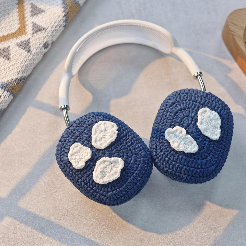 May include: Blue crocheted earmuffs with white crocheted cloud designs. The earmuffs are attached to a white headband.