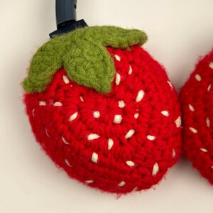 May include: A pair of red crocheted strawberry headphone covers with green leaves and white seeds. The covers are designed to fit over the earcups of headphones. The handmade covers add a playful touch to audio accessories.