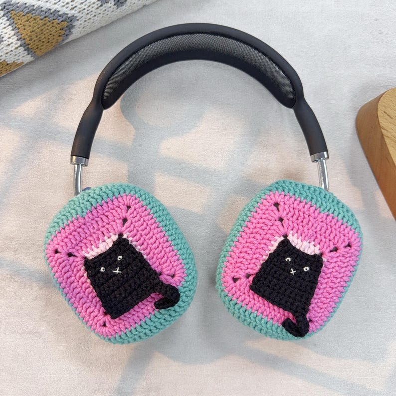 May include: A pair of black headphones with crocheted ear covers. The ear covers are pink, green, and black with a black cat design.