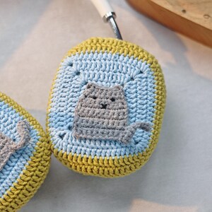 Pu&ograve; includere: Una custodia per cuffie in crochet a forma di gatto, di colore giallo, blu e grigio. La custodia ha un viso di gatto con un naso e baffi grigi.