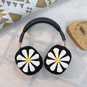 K&ouml;nnte beinhalten: Schwarze Kopfh&ouml;rer mit wei&szlig;en Ohrw&auml;rmern im Daisy-Muster. Die Ohrw&auml;rmer sind geh&auml;kelt und haben eine gelbe Mitte.