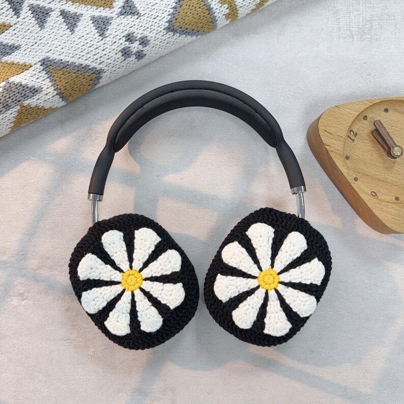 K&ouml;nnte beinhalten: Schwarze Kopfh&ouml;rer mit wei&szlig;en Ohrw&auml;rmern im Daisy-Muster. Die Ohrw&auml;rmer sind geh&auml;kelt und haben eine gelbe Mitte.