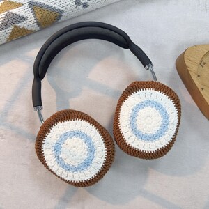 May include: Brown and white crocheted headphone covers with a blue circle design. The covers are attached to a black headband.