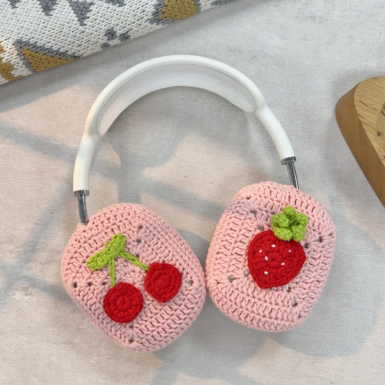 May include: Pink crocheted headphone covers with a strawberry and cherry design. The covers are made of soft yarn and feature a detailed embroidered design.