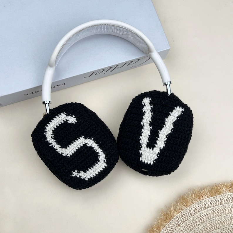 Puede incluir: Auriculares blancos y plateados con almohadillas negras. Las almohadillas est&aacute;n tejidas con las letras "S" y "V" en blanco. Los auriculares descansan sobre una superficie de color claro.