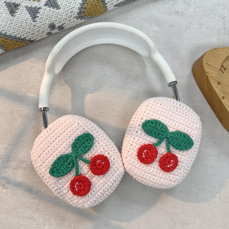 May include: Pink crocheted headphone covers with a cherry design. The cherries are red with green leaves.