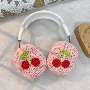 K&ouml;nnte beinhalten: Rosa gestrickte Kopfh&ouml;rerabdeckungen mit einem Kirschendesign. Die Abdeckungen sind aus weichem Garn gefertigt und zeigen ein Paar rote Kirschen mit gr&uuml;nen Stielen und Bl&auml;ttern.