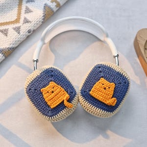 May include: White headphones with crocheted earmuffs featuring a cute orange cat design. The earmuffs are blue and beige with a light brown outline.
