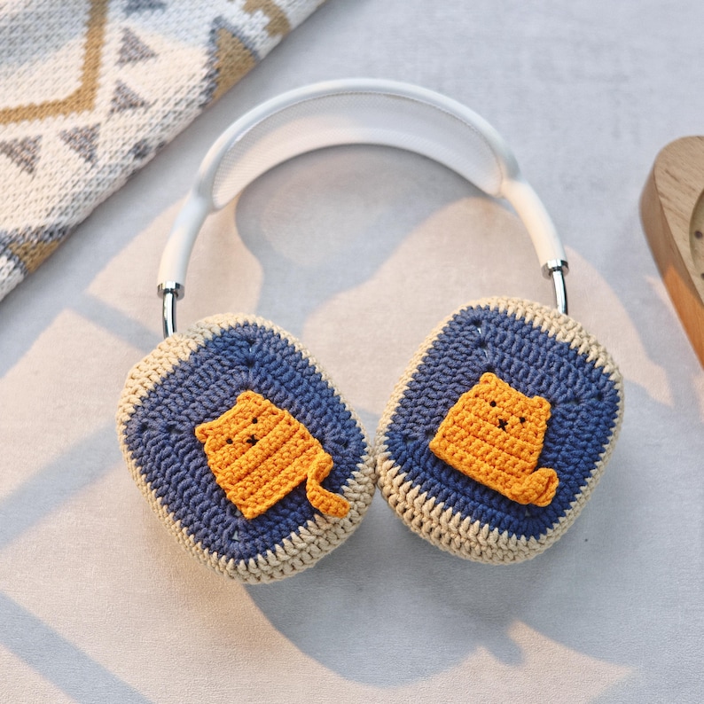 May include: White headphones with crocheted earmuffs featuring a cute orange cat design. The earmuffs are blue and beige with a light brown outline.