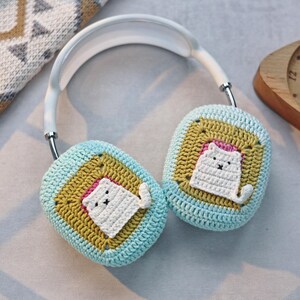 May include: A pair of white headphones with crocheted ear covers. The ear covers are blue and green with a yellow square in the center. Each square has a white crocheted cat sitting inside.