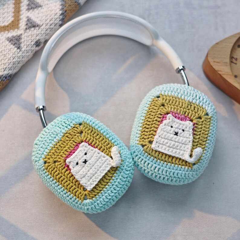 May include: A pair of white headphones with crocheted ear covers. The ear covers are blue and green with a yellow square in the center. Each square has a white crocheted cat sitting inside.