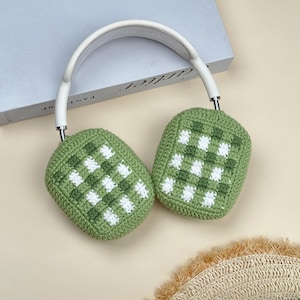 May include: White and silver headphones with green and white checkered crochet ear covers. The headphones rest on a light-colored surface, with a book visible in the background. The ear covers are a unique, handmade design.