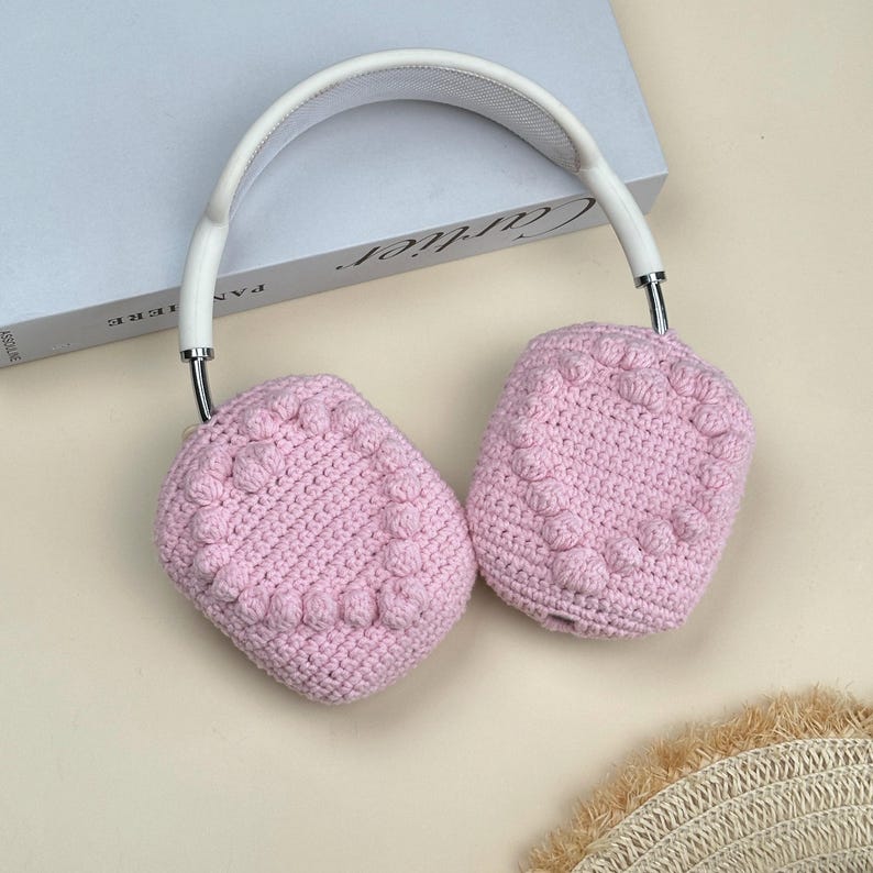 Op de afbeelding: Roze gehaakte hoofdtelefoonhoezen met een textuur van bobbelsteken. De hoezen zijn over witte hoofdtelefoons met zilverkleurige metalen accenten geplaatst. De hoofdtelefoon ligt op een lichtgekleurd oppervlak.
