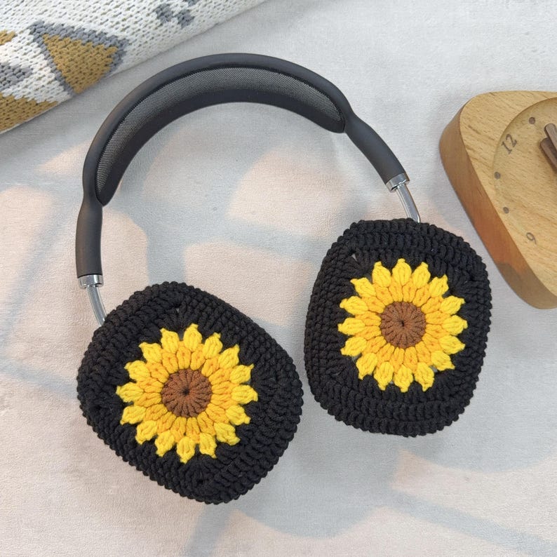 May include: Black headphones with crocheted sunflower ear covers. The ear covers are black with a yellow sunflower in the center. The sunflower has a brown center.