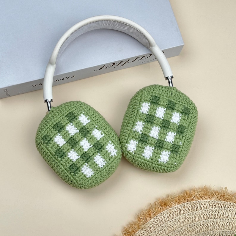 May include: White and silver headphones with green and white checkered crochet ear covers. The headphones rest on a light-colored surface, with a book visible in the background.
