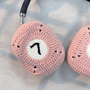 K&ouml;nnte beinhalten: Eine rosa geh&auml;kelte Kopfh&ouml;rerh&uuml;lle mit einem wei&szlig;en Kreis und der Zahl "7" in schwarzer Stickerei.