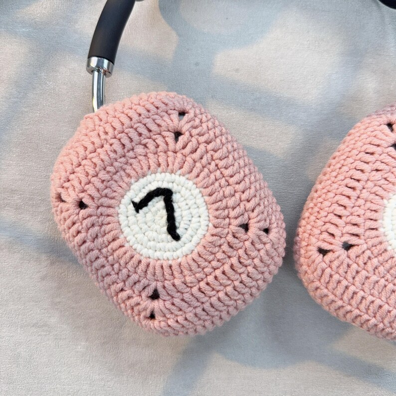 K&ouml;nnte beinhalten: Eine rosa geh&auml;kelte Kopfh&ouml;rerh&uuml;lle mit einem wei&szlig;en Kreis und der Zahl "7" in schwarzer Stickerei.