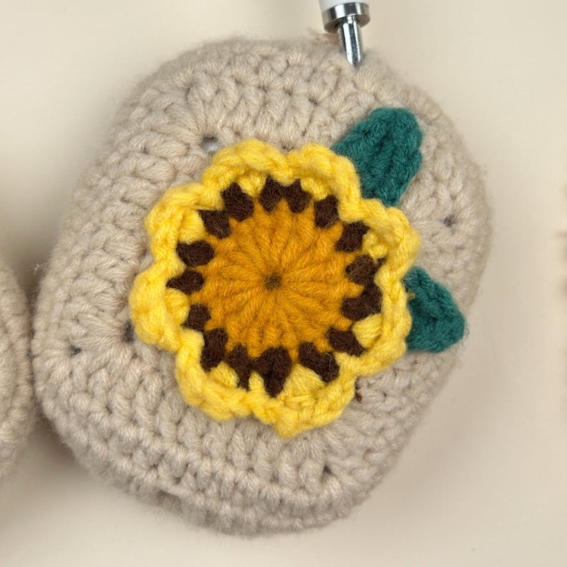 Op de afbeelding: Gehaakte hoofdtelefoonhoes in lichtbeige, versierd met een felgele en bruine zonnebloem en groene bladeren. Het handgemaakte ontwerp voegt een vleugje artisticiteit toe aan de hoofdtelefoon en biedt zowel stijl als bescherming.
