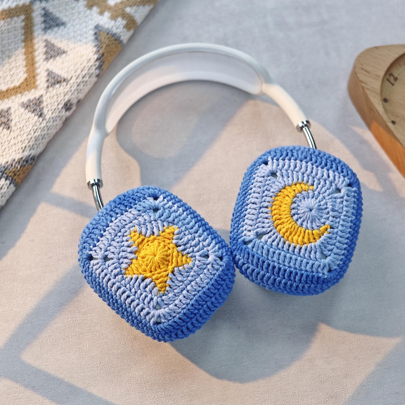 Puede incluir: Orejeras de crochet azul con un dise&ntilde;o de estrella amarilla y media luna. Las orejeras est&aacute;n unidas a una diadema blanca.