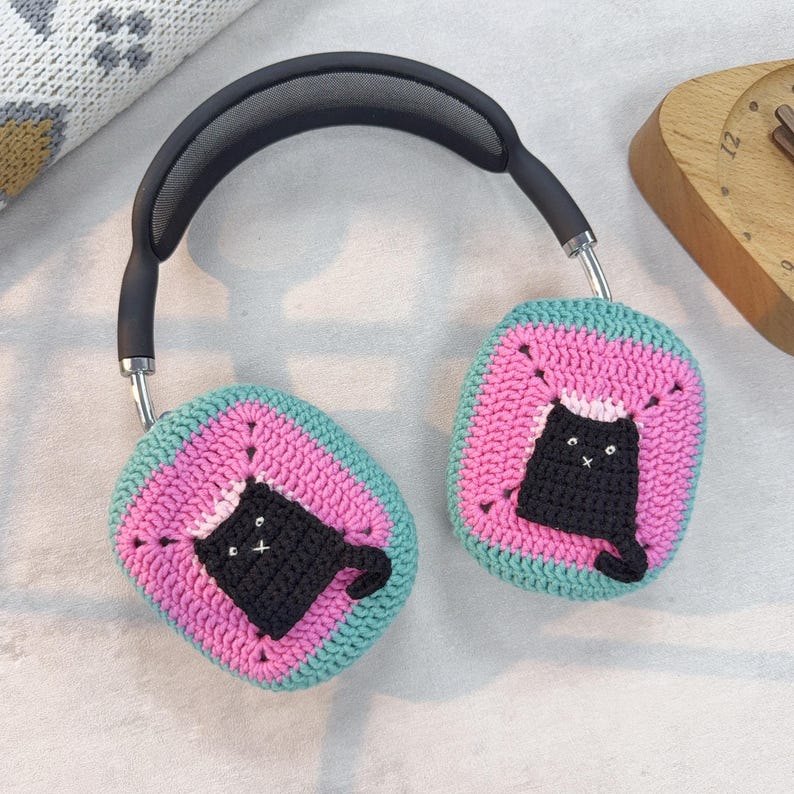 May include: A pair of black headphones with crocheted earmuffs. The earmuffs are pink and teal with a black crocheted cat design.