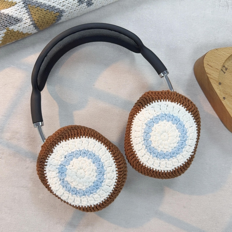 May include: Brown and white crocheted headphone covers with a blue and white circle pattern. The covers are on a pair of black headphones.