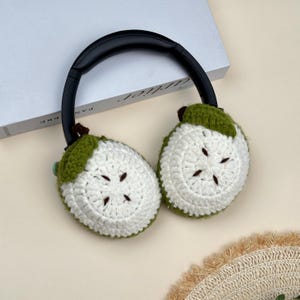 Puede incluir: Auriculares negros con cubiertas para los o&iacute;dos de ganchillo blancas y verdes. Las cubiertas para los o&iacute;dos est&aacute;n dise&ntilde;adas para parecerse a fruta en rodajas, con centros blancos y c&aacute;scaras verdes. Los auriculares est&aacute;n sobre una superficie blanca.