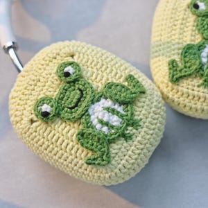 May include: A pair of yellow crocheted headphone covers with green frog appliques. The frogs have white accents and black eyes.