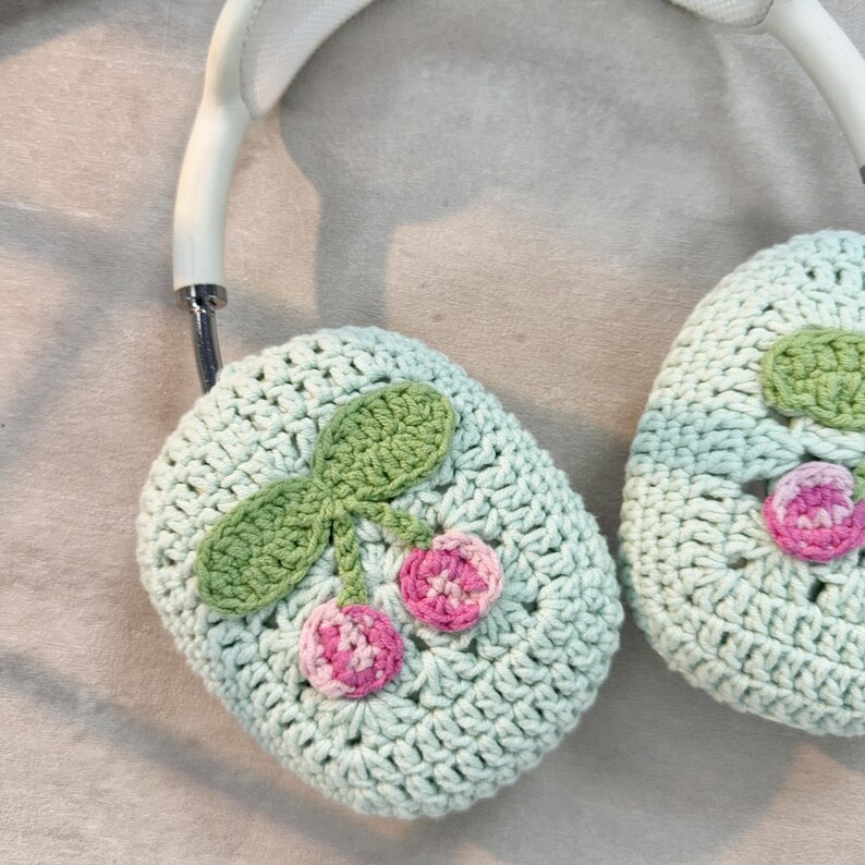 Pu&ograve; includere: Un paio di paraorecchie in crochet verde chiaro con un design a ciliegie. Le ciliegie sono rosa e bianche con foglie verdi.