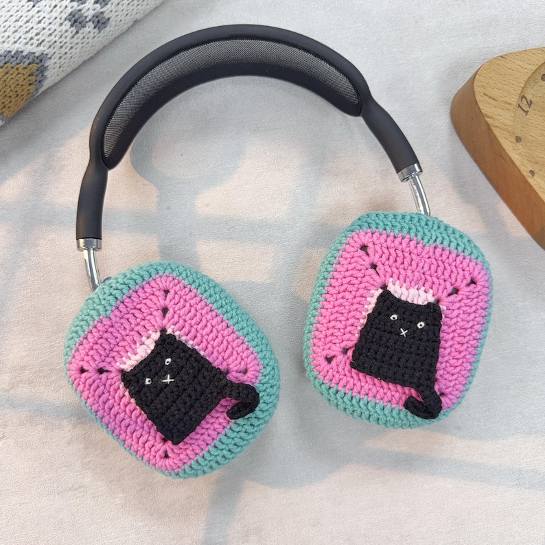 May include: A pair of black headphones with crocheted earmuffs. The earmuffs are pink and green with a black cat design.