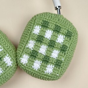 May include: Close-up of a green and white crocheted headphone cover. The cover features a checkerboard pattern with alternating squares of light and dark green, and white floral designs. The headphones are white.
