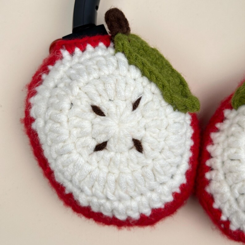 Puede incluir: Fundas de auriculares con forma de manzana tejidas a ganchillo. Las fundas son blancas con un borde rojo, semillas marrones, una hoja verde y un tallo marr&oacute;n. Los auriculares son negros.