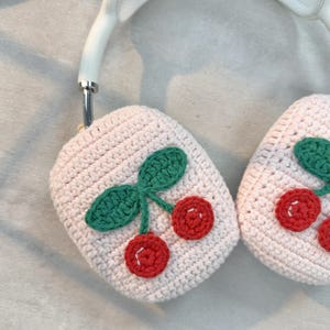 May include: A pair of pink crocheted headphone covers with a cherry design. Each cover features a green crocheted stem with two red cherries.